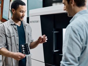 A man holding a clear white part, talking to a another man infront of a CT scanner