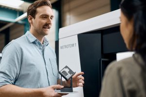 A man holding a clear plastic part, talking to a woman infront of a CT scanner