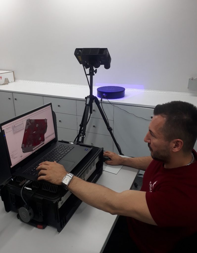 A man setting infront of a laptop, looking at an inspection software. A scan, using a 3D scanner and a rotation table is performed in the background