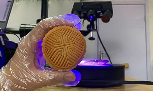 A hand holding a Chinese mooncake in front of the 3D scanner ATOS Q